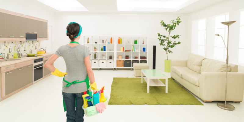 A woman stares at the room that she's about to clean. She's holding cleaning supplies on her right side.