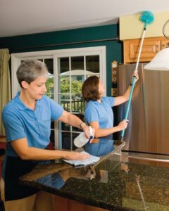 Two house cleaners work on cleaning up the kitchen. The one in the front is spraying and wiping a granite countertop. The one in the back is dusting a hard to reach spot on the ceiling.