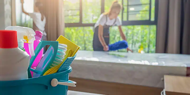 A cleaner stands in the background out of focus as she holds a duster and is dusting.